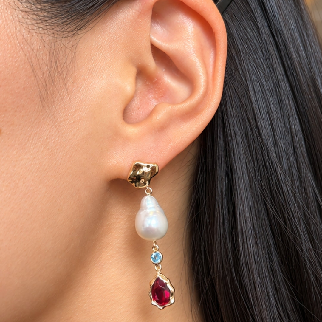 Close-up of a pearl drop earring worn on the ear, featuring a sculptural gold stud, baroque pearl, small round blue gemstone, and pear-shaped red gemstone drop.
