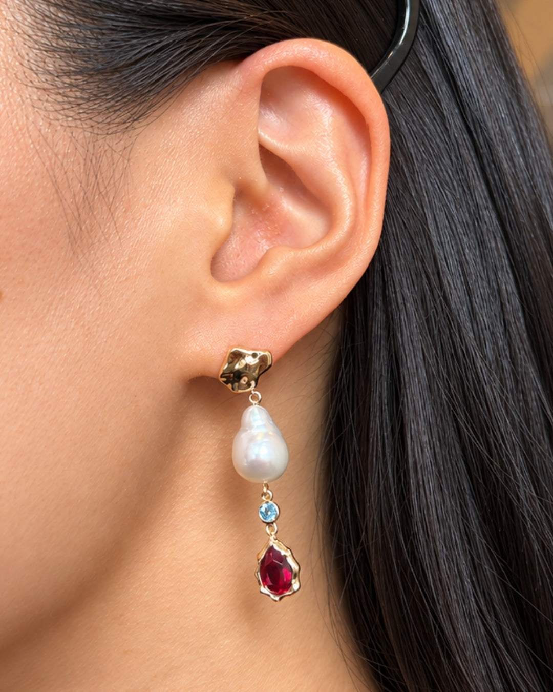 Close-up of a pearl drop earring worn on the ear, featuring a sculptural gold stud, baroque pearl, small round blue gemstone, and pear-shaped red gemstone drop.