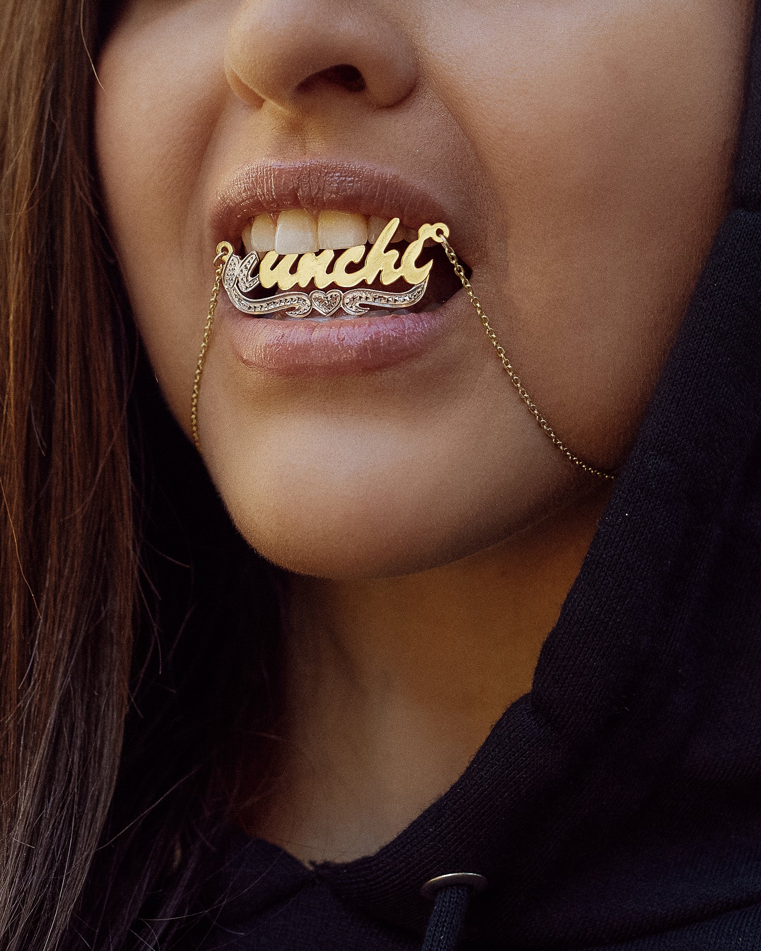 Close-up of a person slightly biting a gold nameplate necklace that spells “nunchi” in cursive lettering, with a diamond-accented underline and heart detail. They wear a black hoodie, and the chain hangs across their lips.