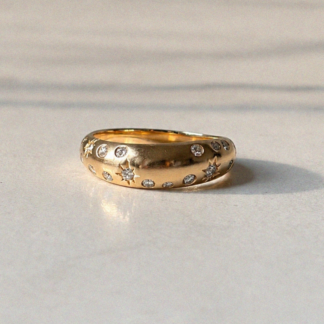 Sculptural polished gold ring with scattered flush-set diamonds and engraved starburst details, photographed in soft sunlight on a pale marble surface.