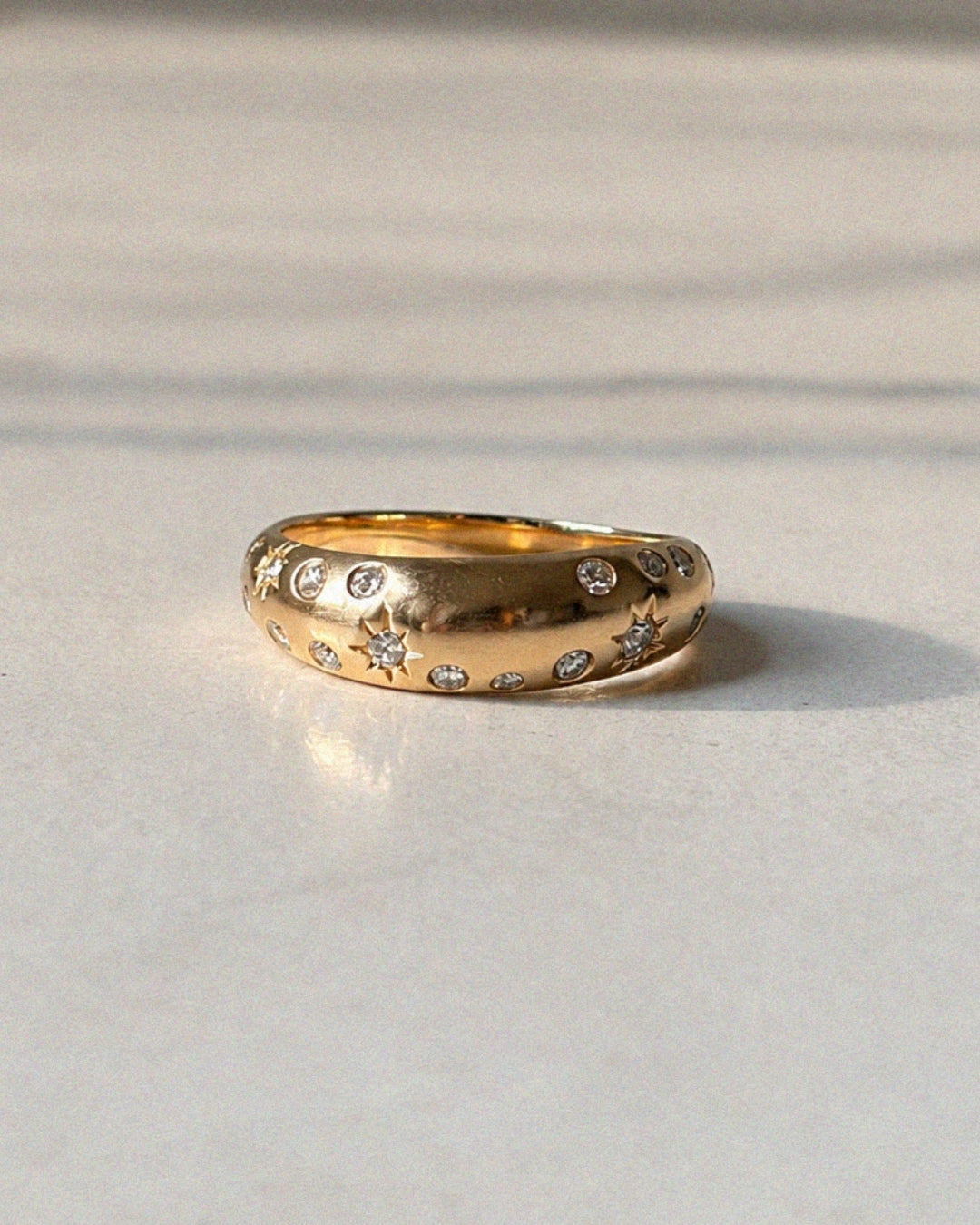 Sculptural polished gold ring with scattered flush-set diamonds and engraved starburst details, photographed in soft sunlight on a pale marble surface.