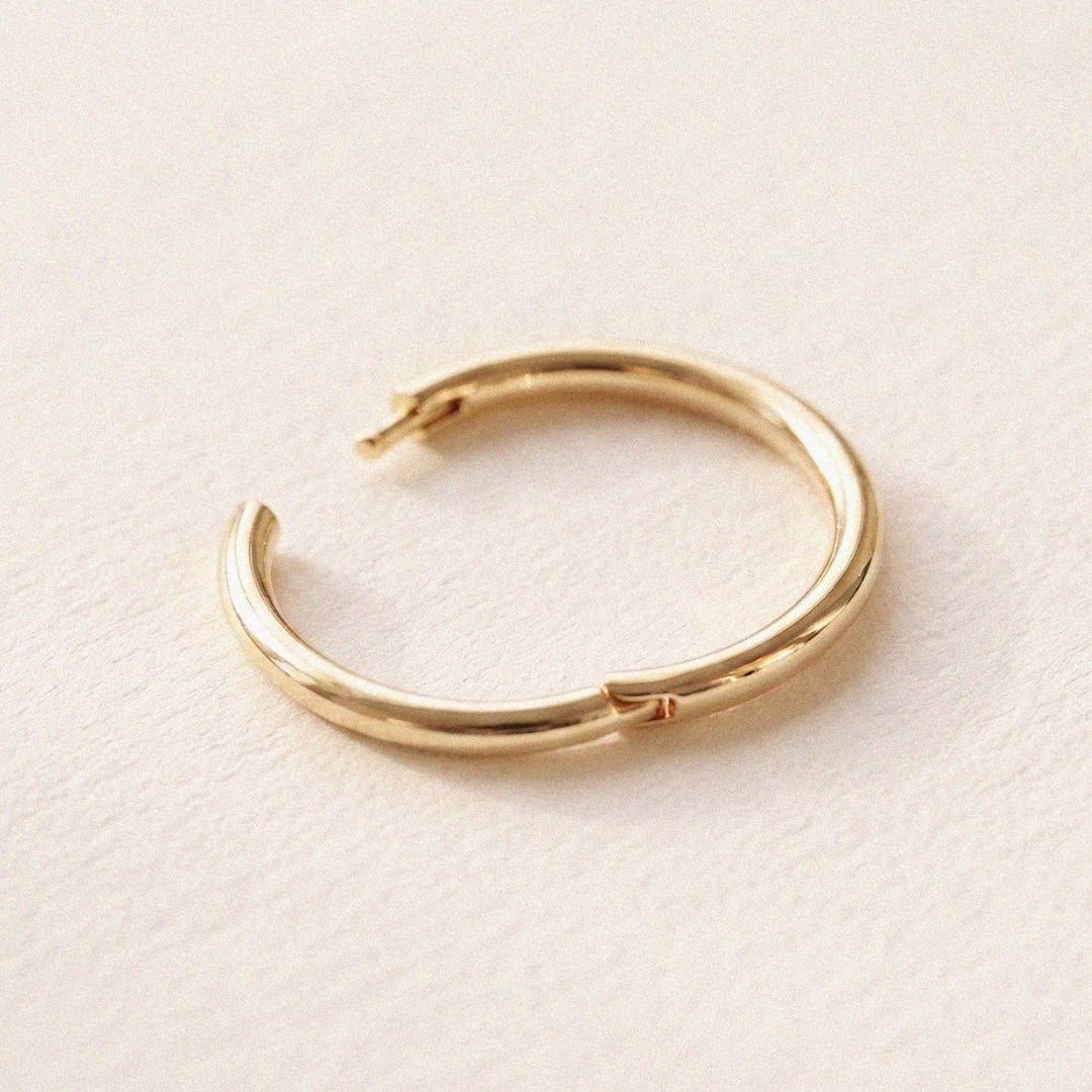 Minimal product photograph of a polished yellow gold hoop earring resting on a textured ivory surface. The hinge closure and smooth rounded silhouette are shown in close detail.