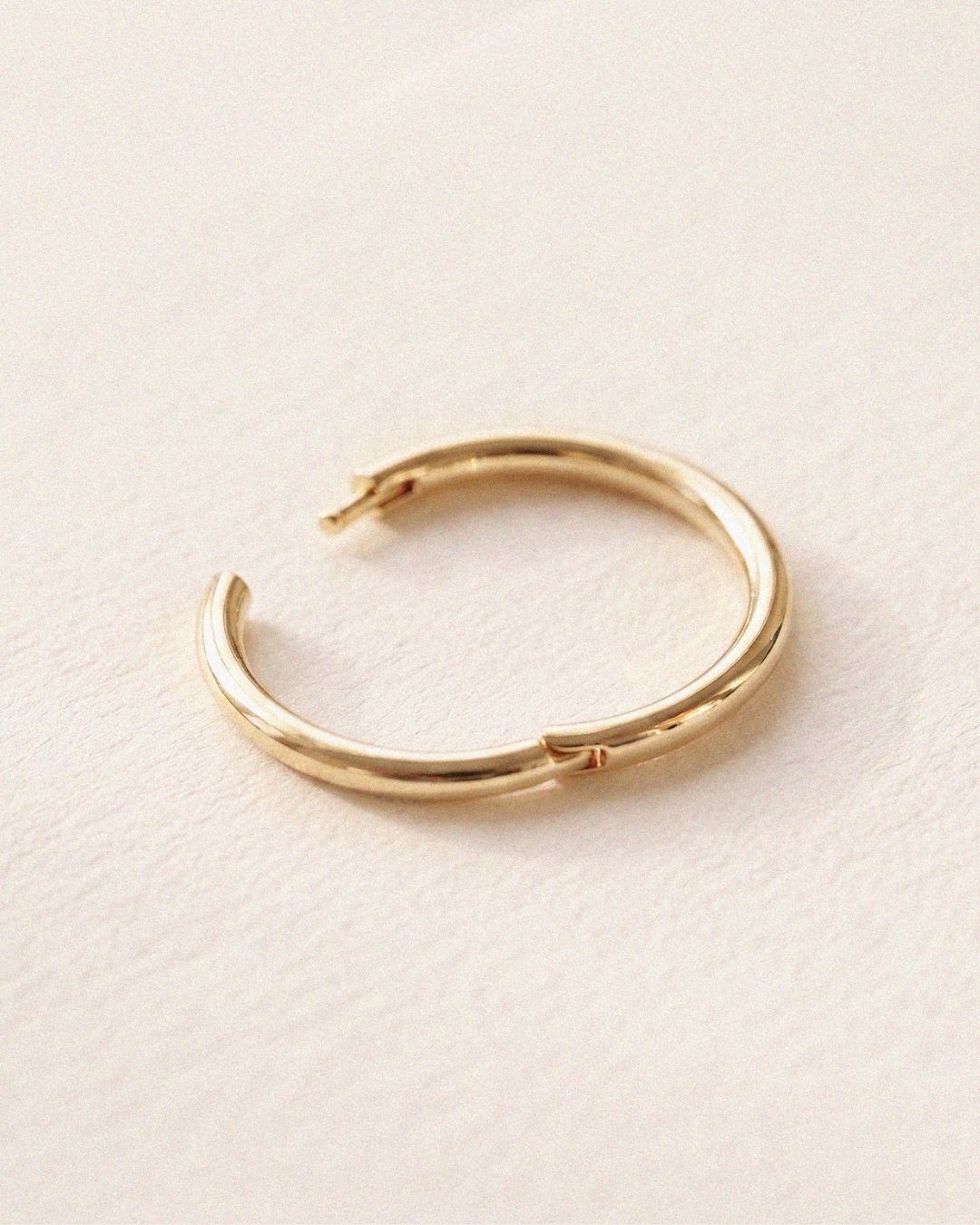 Minimal product photograph of a polished yellow gold hoop earring resting on a textured ivory surface. The hinge closure and smooth rounded silhouette are shown in close detail.