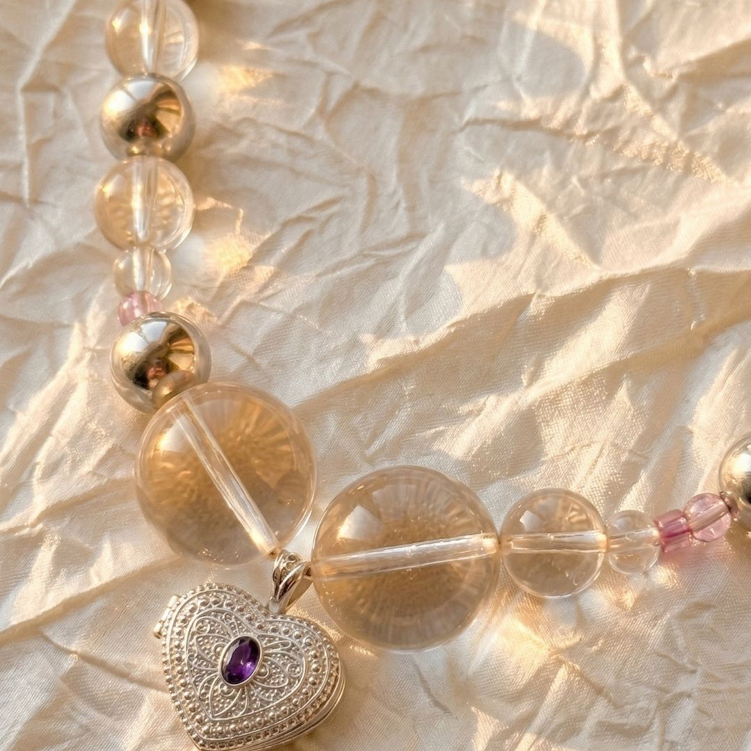 Close-up of a heart-shaped silver locket pendant with a purple gemstone hanging from a necklace made of clear and metallic beads, photographed on softly crinkled fabric in warm sunlight.