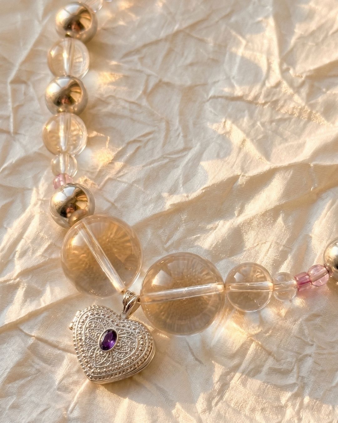 Close-up of a heart-shaped silver locket pendant with a purple gemstone hanging from a necklace made of clear and metallic beads, photographed on softly crinkled fabric in warm sunlight.
