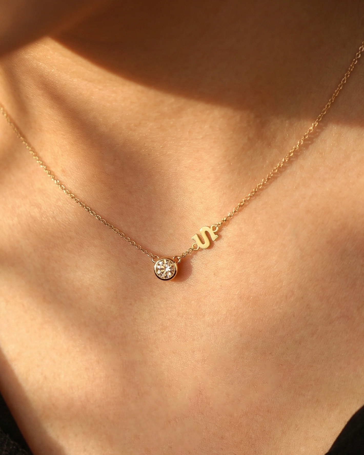 Delicate gold necklace with a bezel-set round diamond pendant and a small initial charm, worn on a model’s neck in warm sunlight.