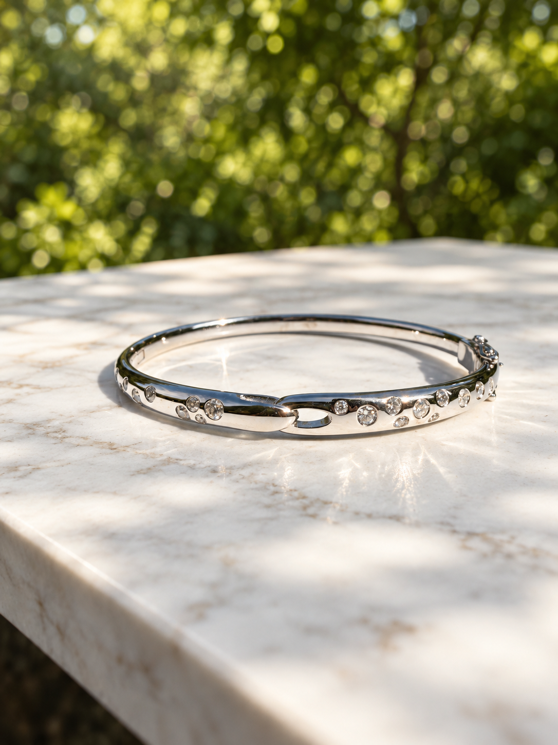 Polished silver hinged bangle bracelet set with scattered round diamonds, photographed outdoors on a white marble surface in direct sunlight.