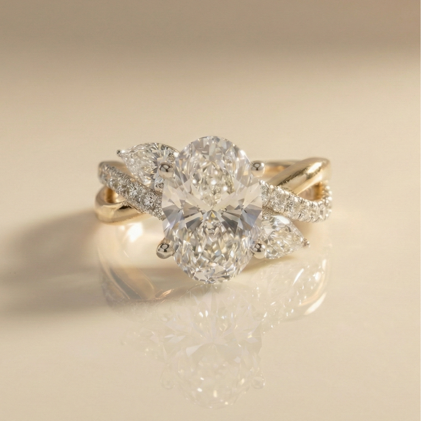 Oval diamond engagement ring with pear-shaped side stones and a twisted pavé band in yellow and white gold, shown on a reflective neutral surface.