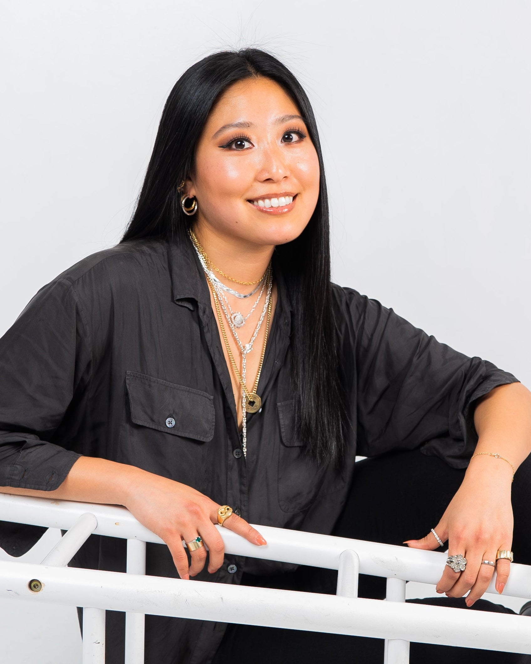 Woman seated on a white chair wearing layered gold necklaces, rings, and hoop earrings against a light gray studio background.