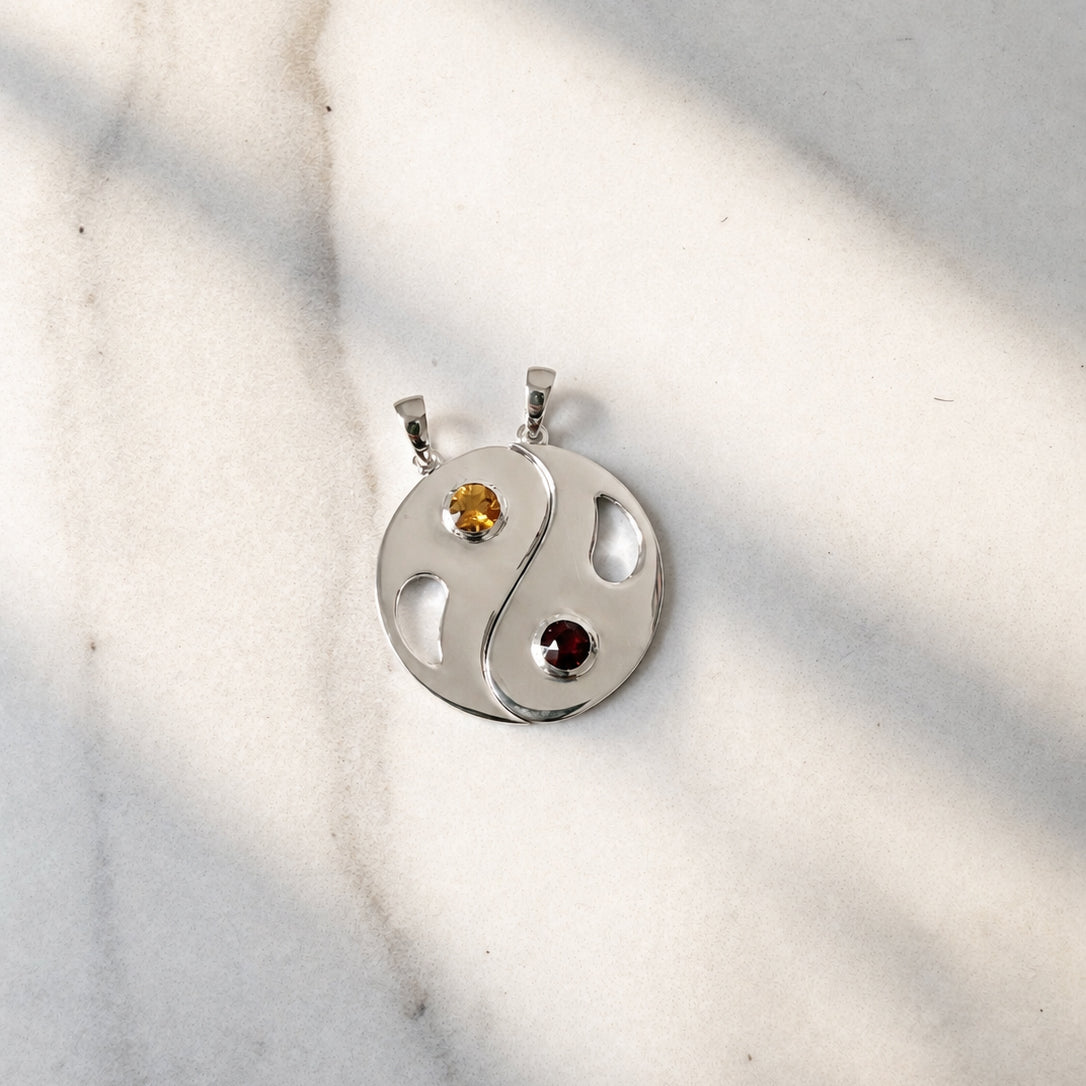 Polished silver Yin Yang pendant set featuring a warm yellow citrine and deep red garnet in round bezel settings, photographed on a softly textured cream surface with natural sunlight and shadows.