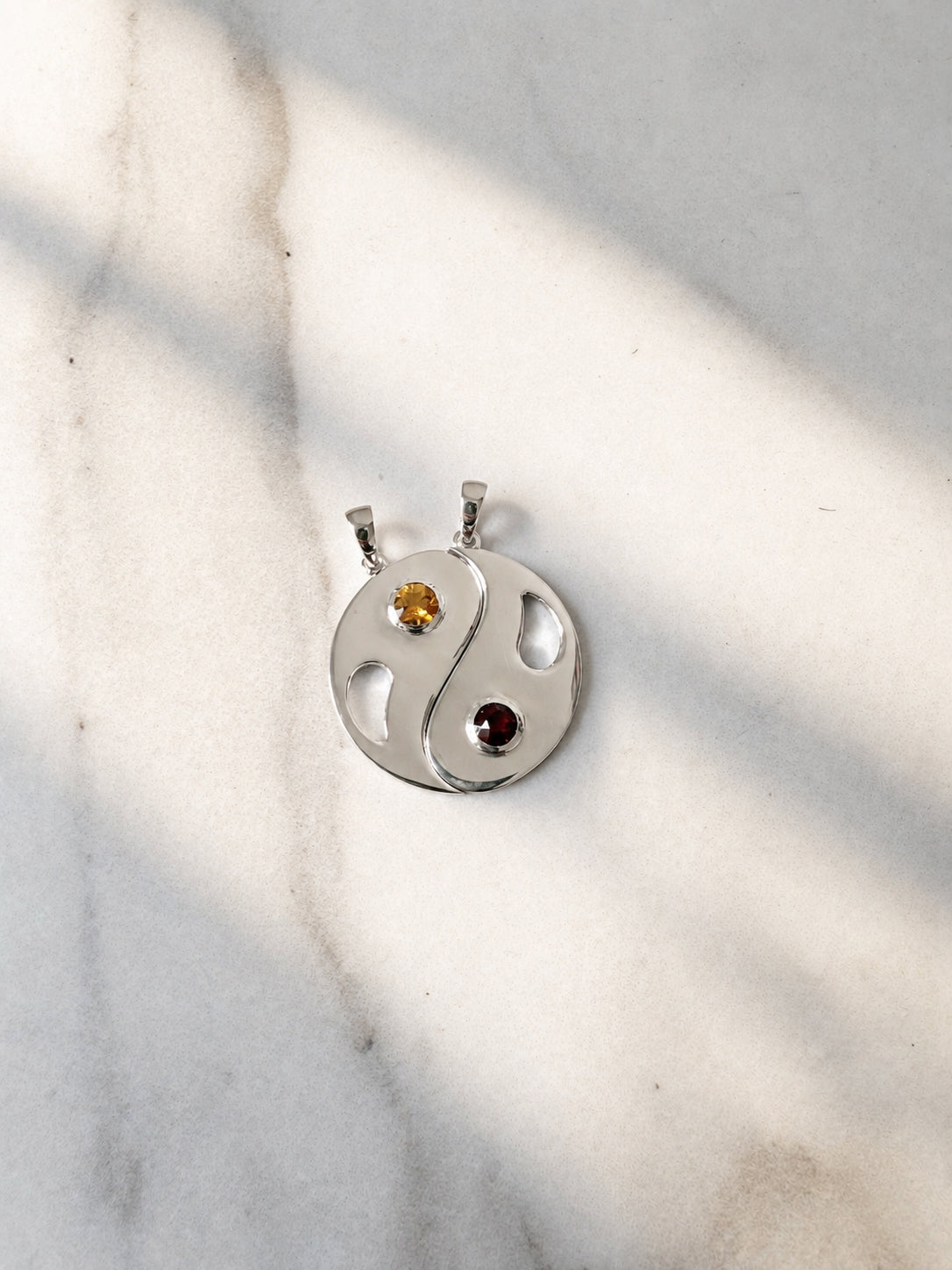 Polished silver Yin Yang pendant set featuring a warm yellow citrine and deep red garnet in round bezel settings, photographed on a softly textured cream surface with natural sunlight and shadows.