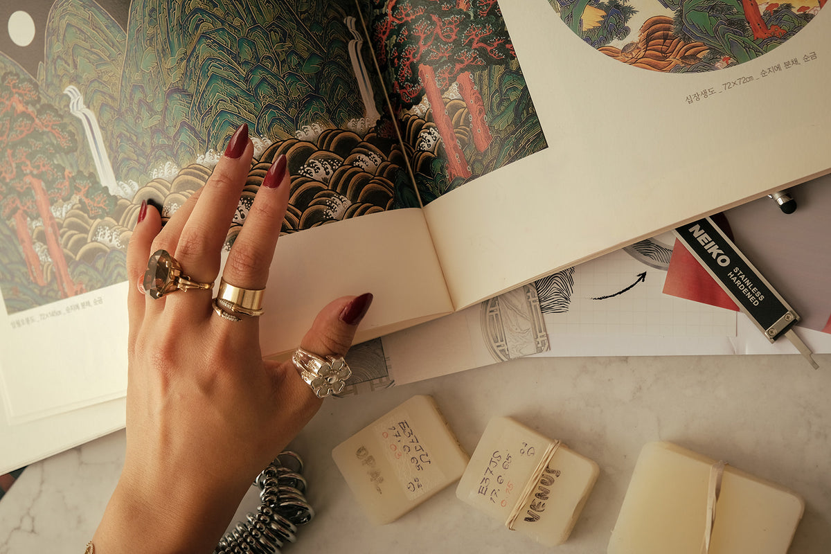 Close-up of Jane Dua’s hand wearing NUNCHI rings beside an open Korean art book, jewelry sketches, measuring tools, and material samples.