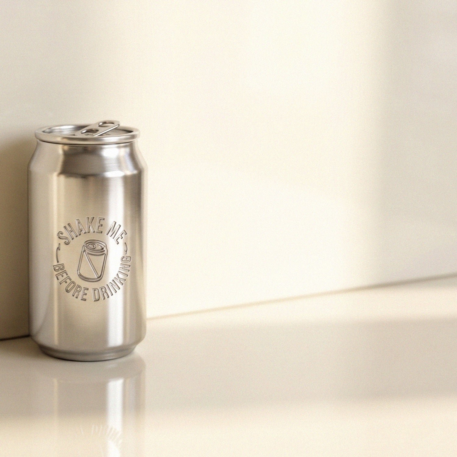 Silver can-shaped pendant engraved with “SHAKE ME BEFORE DRINKING,” photographed on a reflective surface in soft natural light.