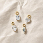Four diamond drop charms in emerald, heart, marquise, and oval cuts, each suspended from a polished gold loop and displayed upright on a warm ivory background.