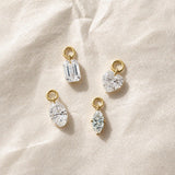 Four diamond drop charms in emerald, heart, marquise, and oval cuts, each suspended from a polished gold loop and displayed upright on a warm ivory background.
