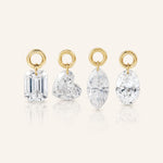 Four diamond drop charms in emerald, heart, marquise, and oval cuts, each suspended from a polished gold loop and displayed upright on a warm ivory background.