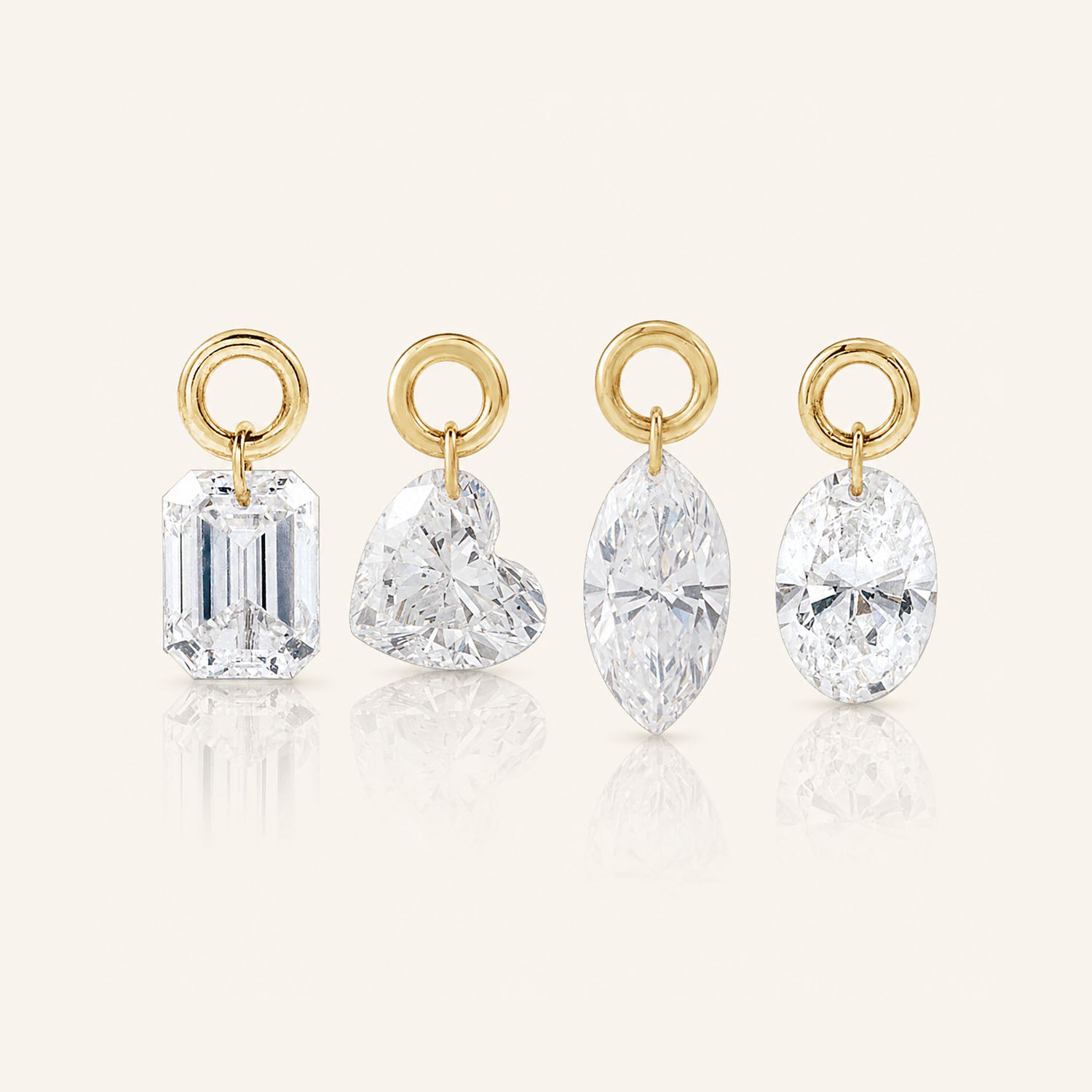 Four diamond drop charms in emerald, heart, marquise, and oval cuts, each suspended from a polished gold loop and displayed upright on a warm ivory background.