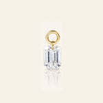 Emerald-cut diamond drop charm suspended from a polished gold loop, photographed with a subtle reflection on a warm ivory background.
