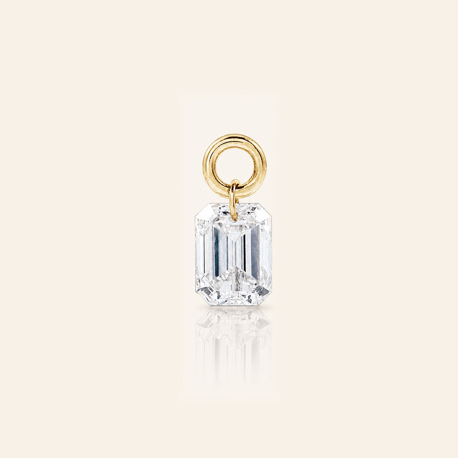Emerald-cut diamond drop charm suspended from a polished gold loop, photographed with a subtle reflection on a warm ivory background.