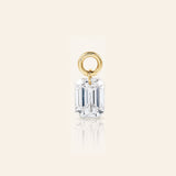 Emerald-cut diamond drop charm suspended from a polished gold loop, photographed with a subtle reflection on a warm ivory background.