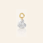 Heart-shaped diamond drop charm suspended from a polished gold loop, photographed with a subtle reflection on a warm ivory background.