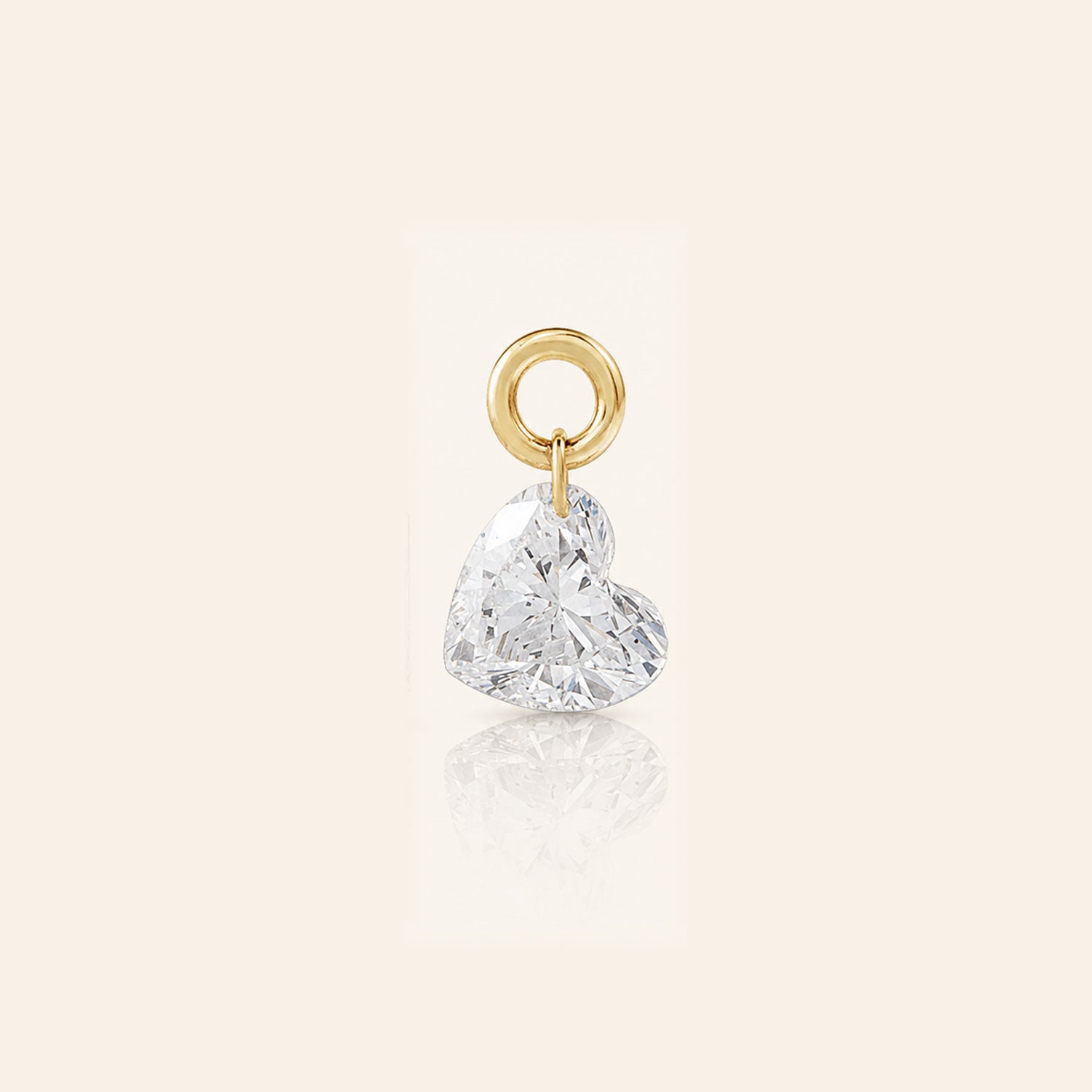 Heart-shaped diamond drop charm suspended from a polished gold loop, photographed with a subtle reflection on a warm ivory background.
