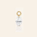 Oval-cut diamond drop charm suspended from a polished gold loop, photographed with a subtle reflection on a warm ivory background.