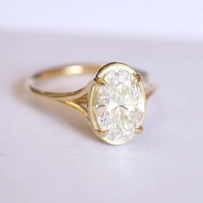 Oval diamond solitaire engagement ring set in yellow gold with a milgrain bezel, shown on a clean white background.