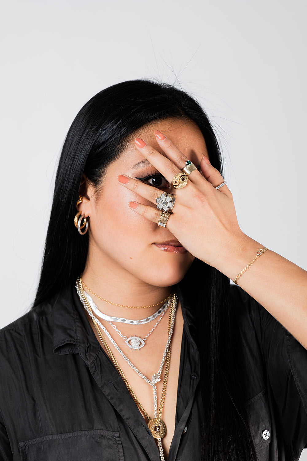 NUNCHI Founder Jane Dua wearing layered gold and silver jewelry including statement rings, hoop earrings, and pendant necklaces, covering one eye with her hand.