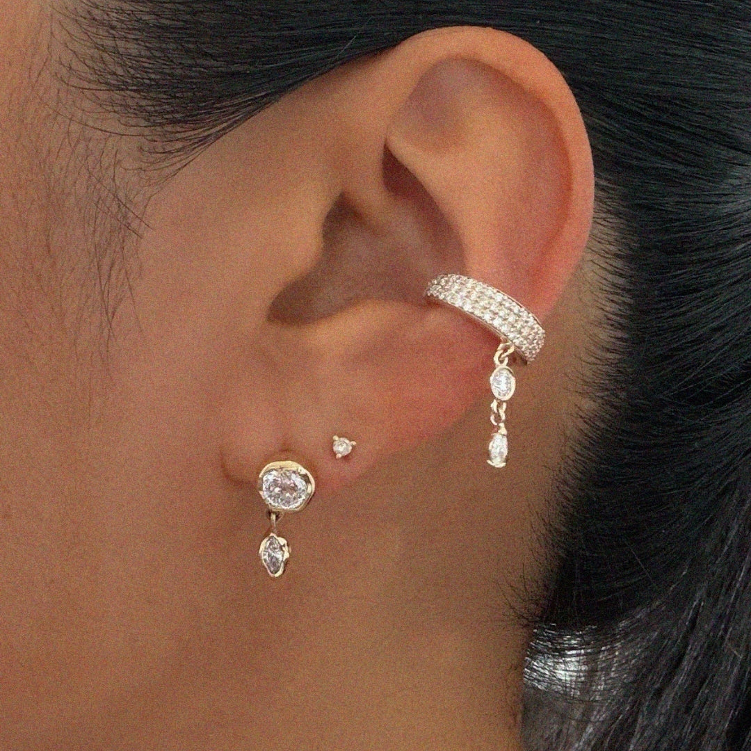 Close-up of an ear styled with multiple diamond earrings. A yellow gold bezel-set diamond stud with a marquise diamond drop is paired with a small heart-shaped diamond stud and a pavé diamond ear cuff featuring a cascading diamond charm. The jewelry is shown on a model with dark hair pulled back, highlighting the layered ear stack.