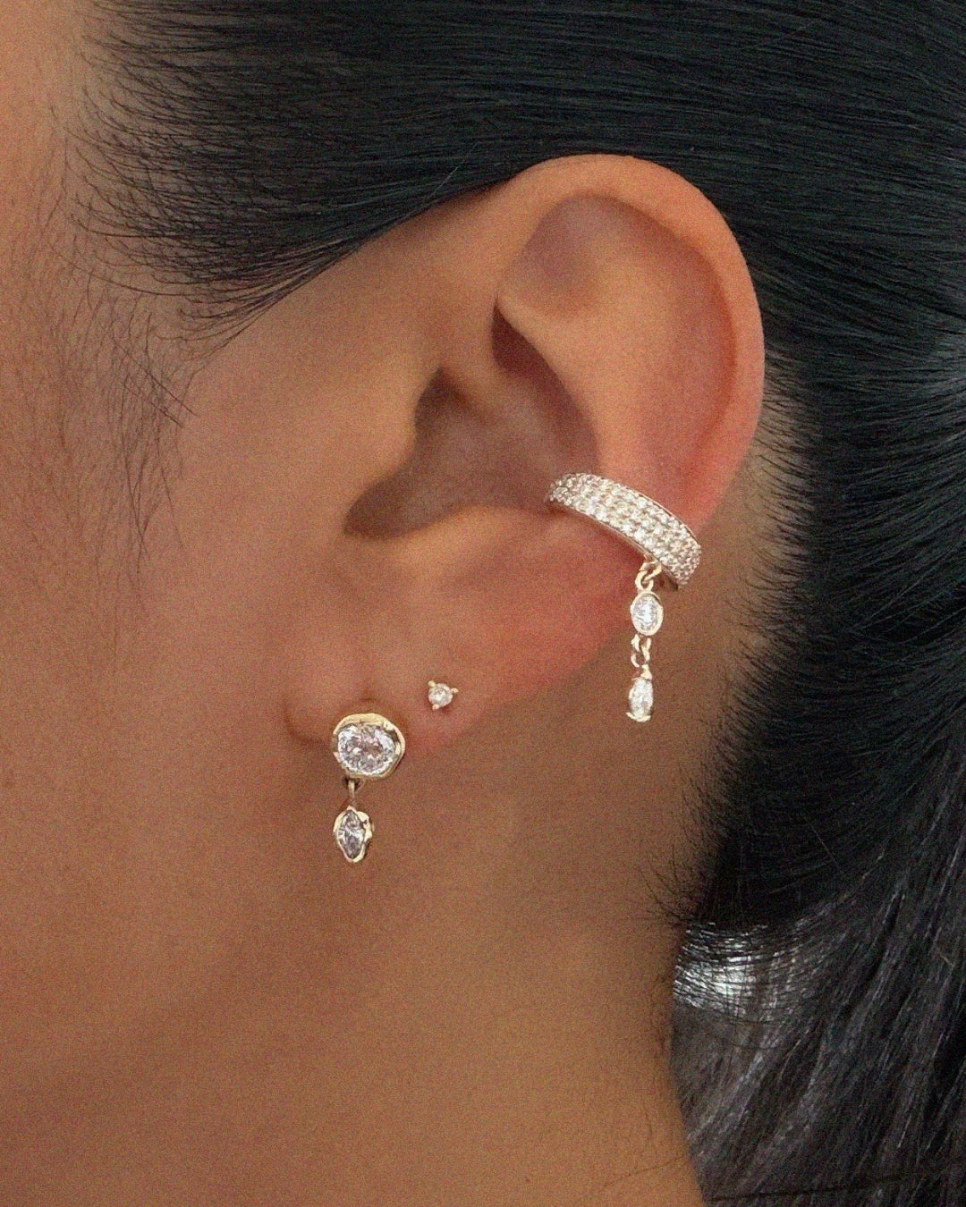 Close-up of an ear styled with multiple diamond earrings. A yellow gold bezel-set diamond stud with a marquise diamond drop is paired with a small heart-shaped diamond stud and a pavé diamond ear cuff featuring a cascading diamond charm. The jewelry is shown on a model with dark hair pulled back, highlighting the layered ear stack.
