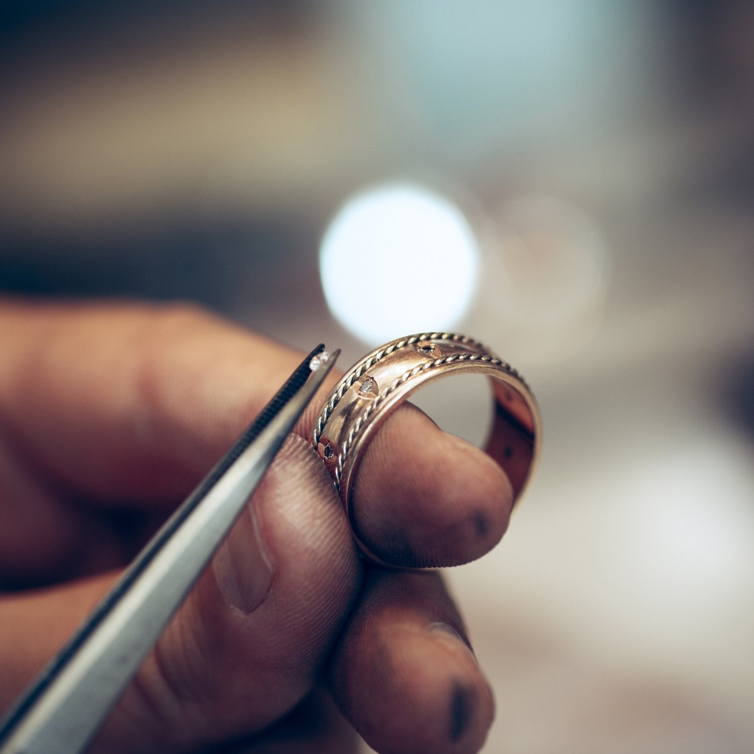 Close-up of a handcrafted gold band with milgrain edges and small diamond accents, held in tweezers during the jewelry setting process.