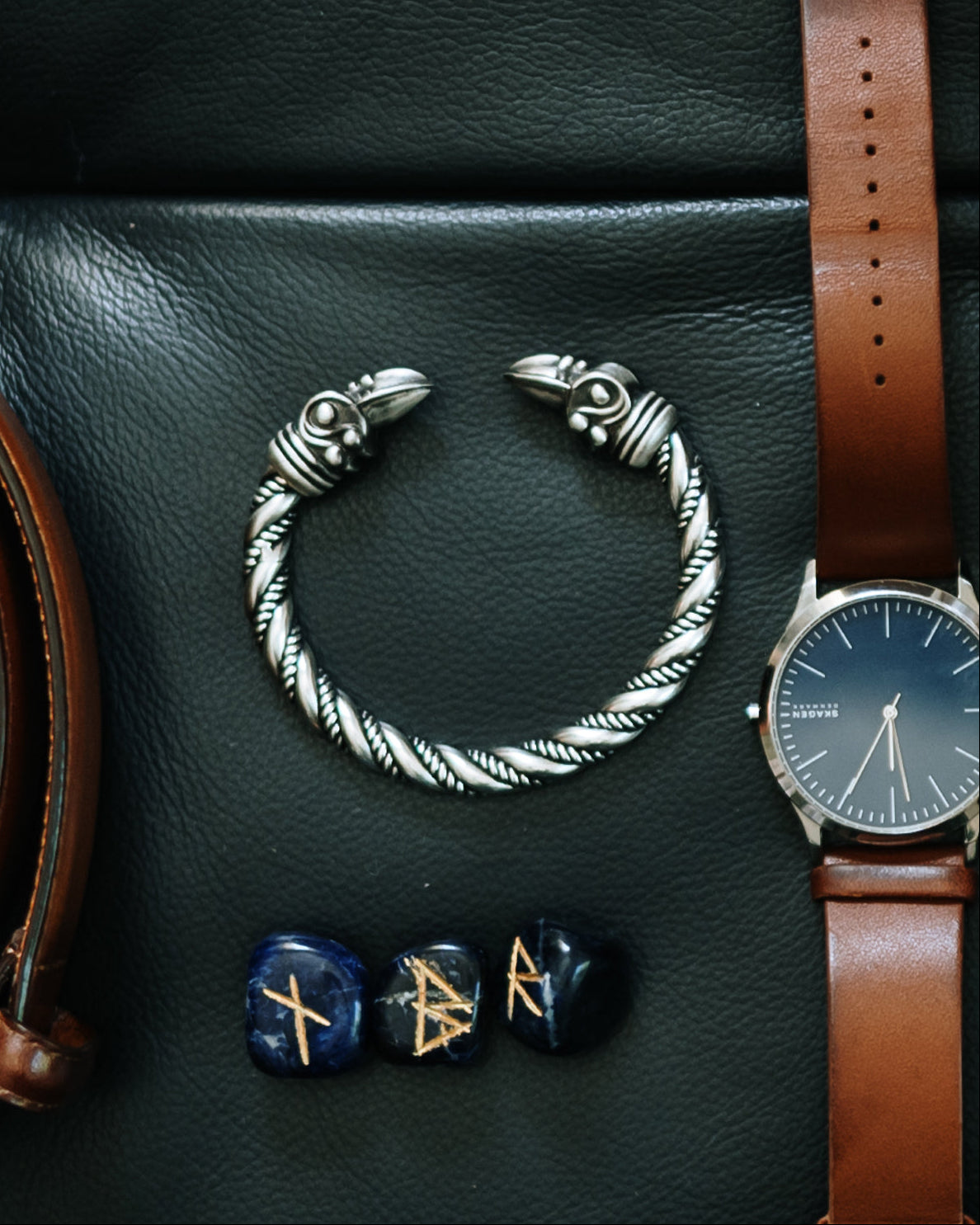 Twisted metal raven head bracelet placed on dark leather beside a brown leather watch strap and blue rune stones.