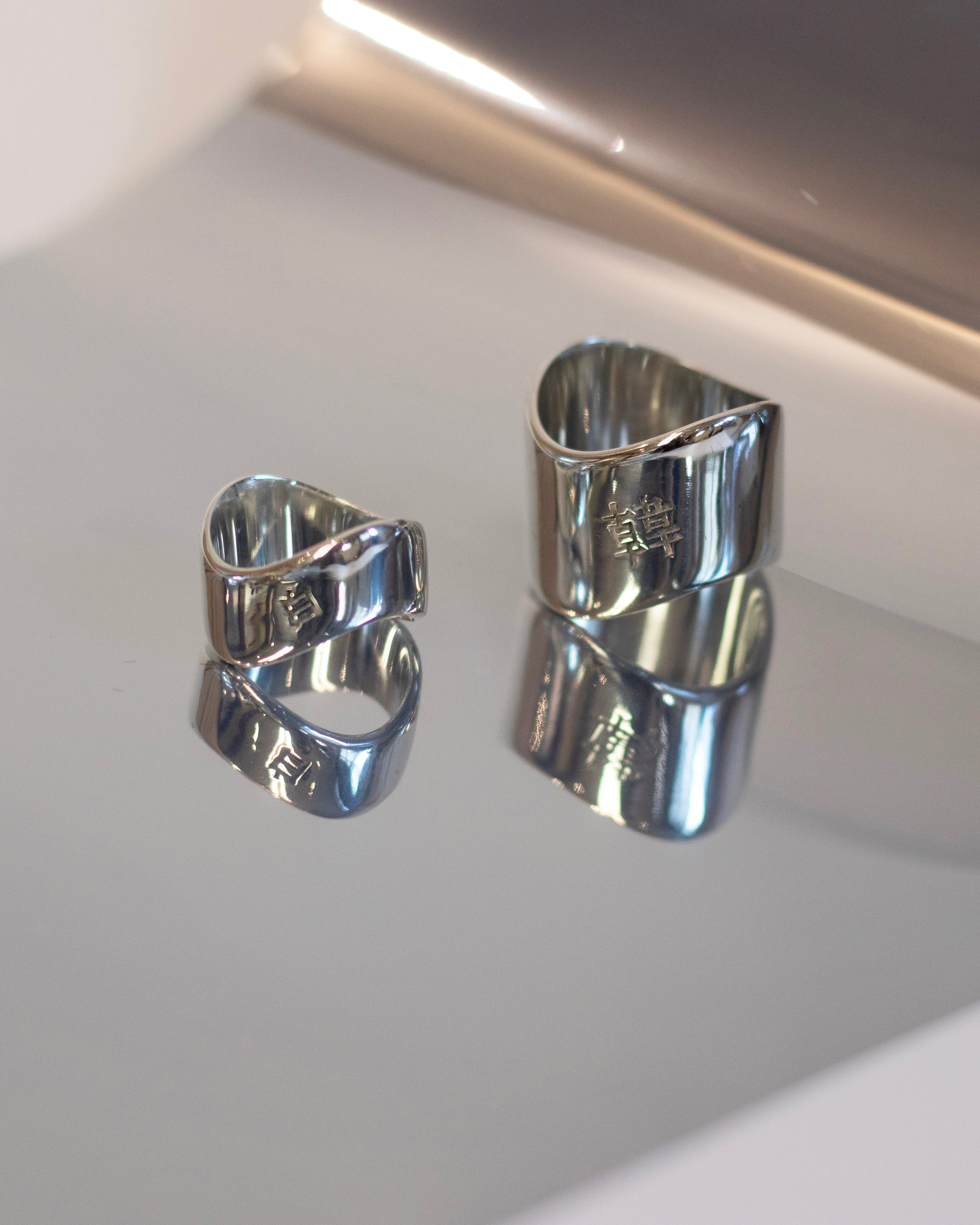 Two polished silver sculptural band rings with engraved Chinese characters displayed on a reflective surface.