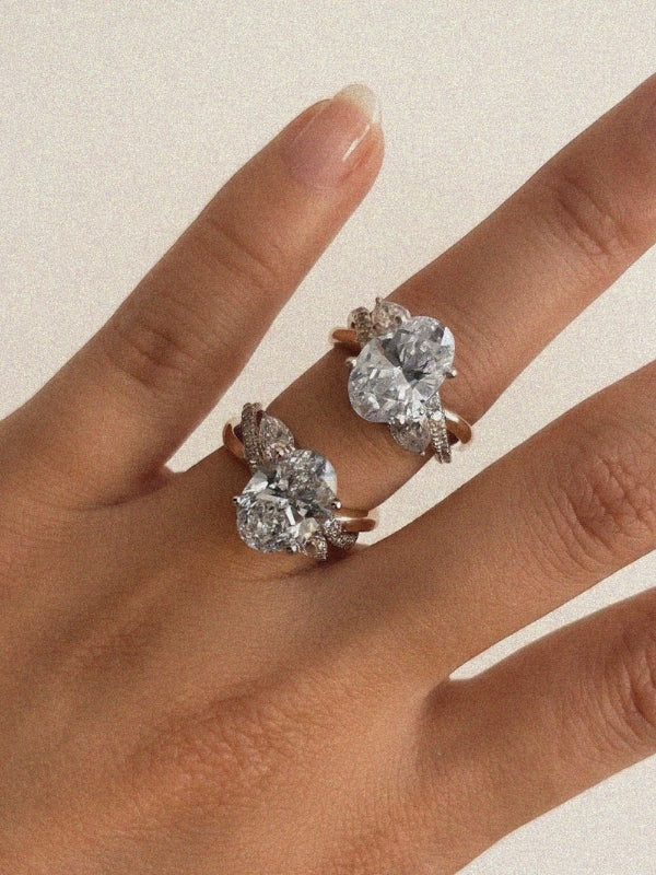 Close-up of a hand wearing two diamond rings on adjacent fingers. Each ring features a large oval center stone with pear-shaped and pavé diamond accents set on yellow gold bands, photographed against a soft neutral background in warm natural light.