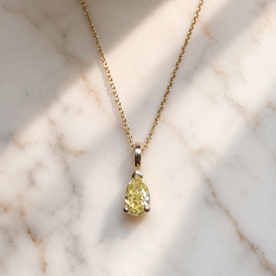 Close-up of a pear-shaped fancy yellow diamond pendant suspended from a delicate yellow gold chain. The diamond is secured in a minimalist three-prong setting with a polished bail and photographed on white marble in soft natural sunlight, emphasizing the stone's warm golden hue and brilliance.