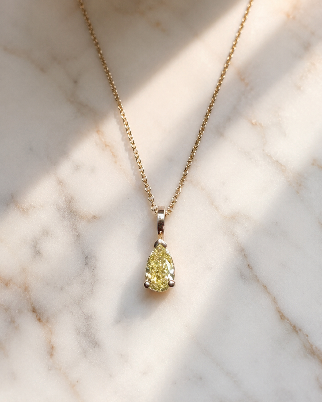 Close-up of a pear-shaped fancy yellow diamond pendant suspended from a delicate yellow gold chain. The diamond is secured in a minimalist three-prong setting with a polished bail and photographed on white marble in soft natural sunlight, emphasizing the stone's warm golden hue and brilliance.