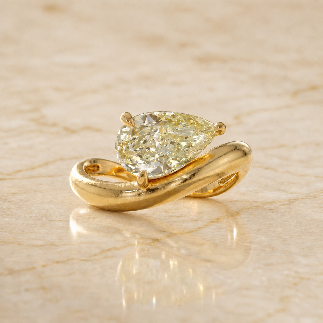 Pear-shaped diamond solitaire set in a sculptural, high-polish gold band with soft curves, photographed in warm sunlight on a marble surface with a subtle reflection.
