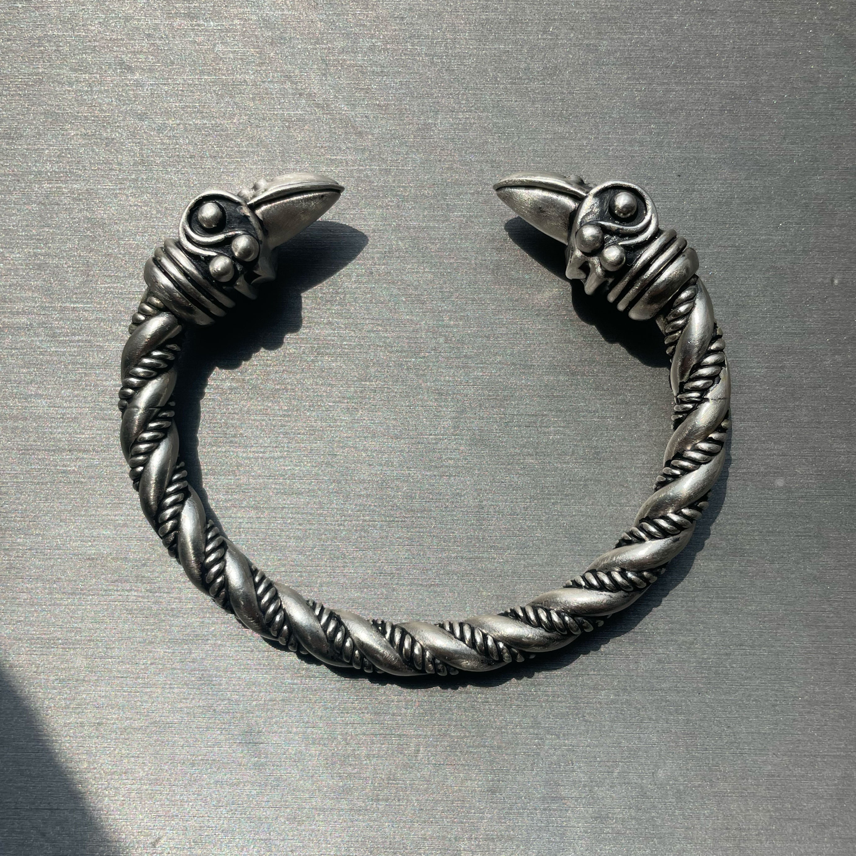 Oxidized silver twisted cuff bracelet with detailed raven-head ends, photographed on a brushed metal surface.