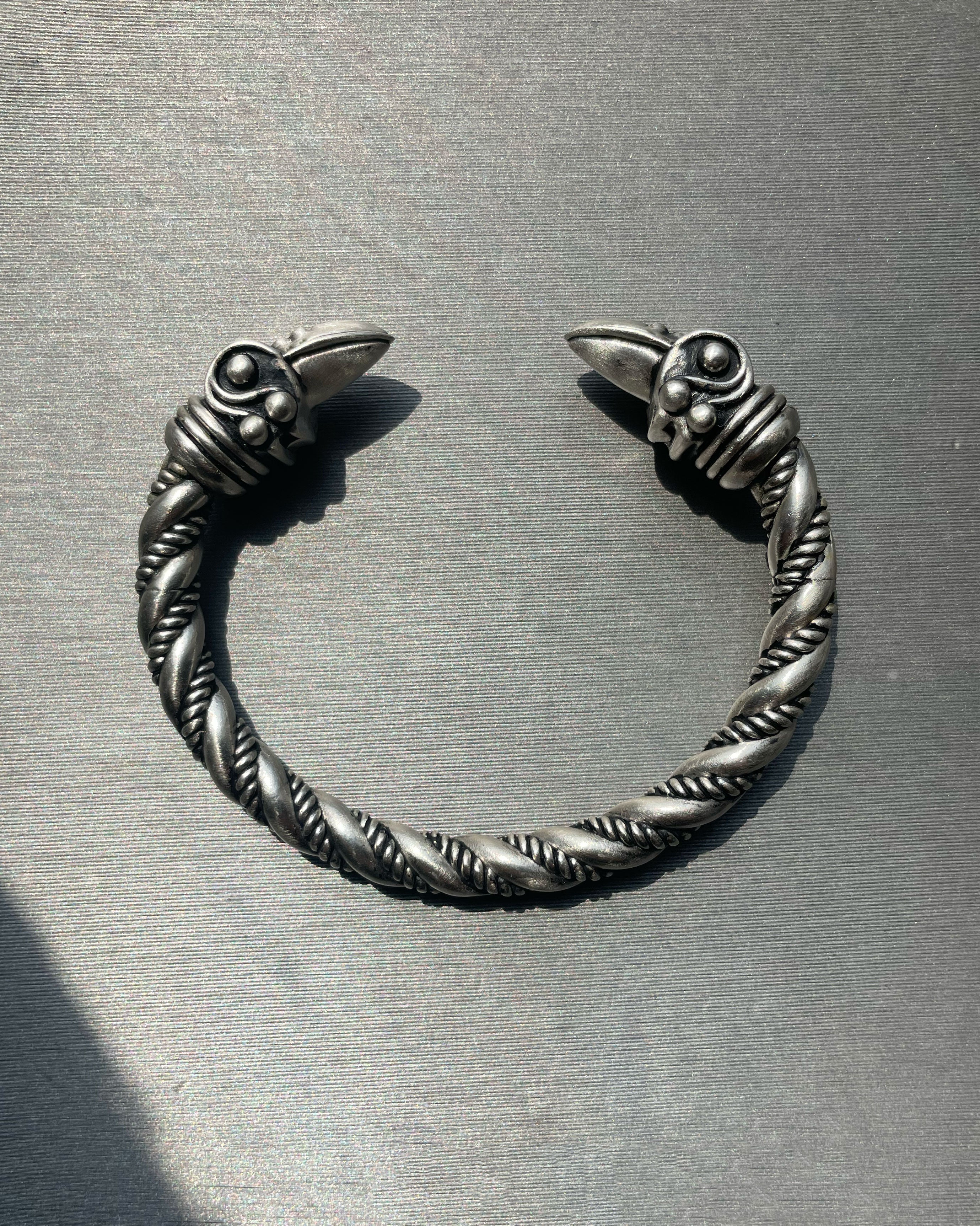 Oxidized silver twisted cuff bracelet with detailed raven-head ends, photographed on a brushed metal surface.